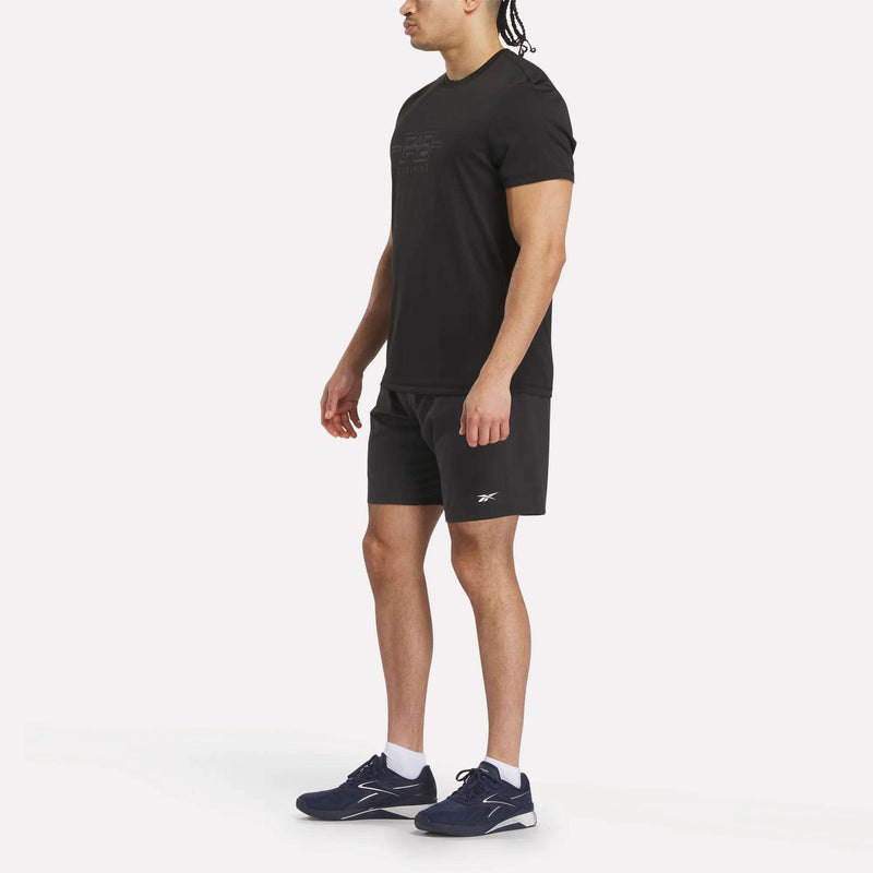 A man stands against a white background, wearing the Men’s Reebok x F45 Training T-Shirt, black shorts, white socks, and navy blue Reebok athletic shoes. He faces slightly left with relaxed arms—ideal for F45 Training or casual wear.