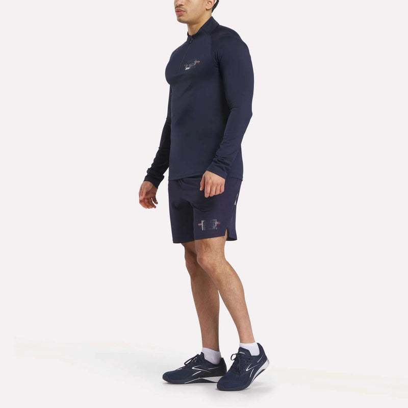 A man in a Reebok x F45 Training Coach Quarter-Zip Top, black shorts, and navy running shoes stands against a plain white background, facing slightly to the side.
