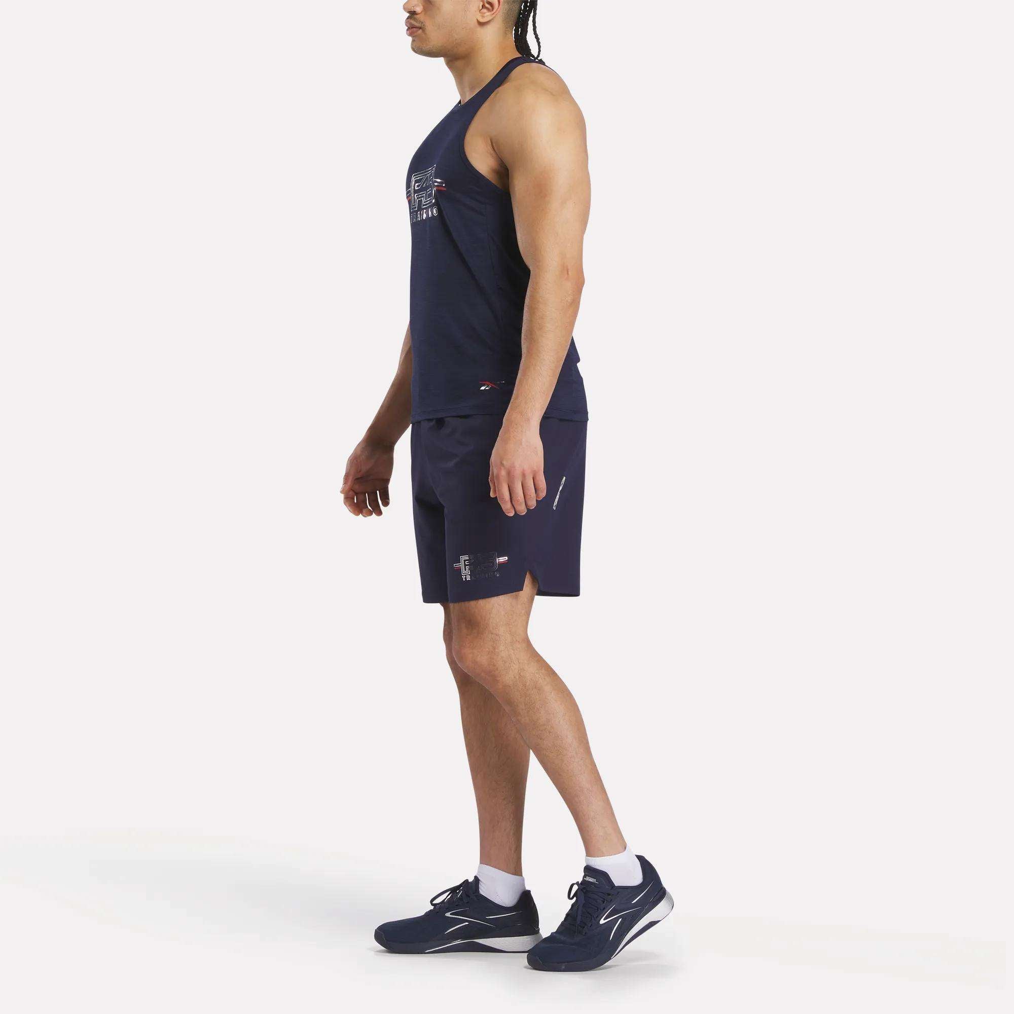 A man stands sideways on a plain white background wearing a Men's Reebok x F45 Training Coach Tank Top, matching navy shorts, white socks, and navy running shoes.