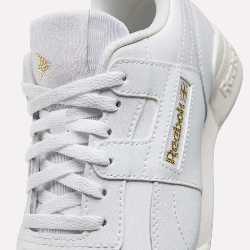A close-up of Reebok Workout Plus Shoes for big kids, showcasing white textured leather, gold Reebok logos on the tongue and side, and crisp white laces.