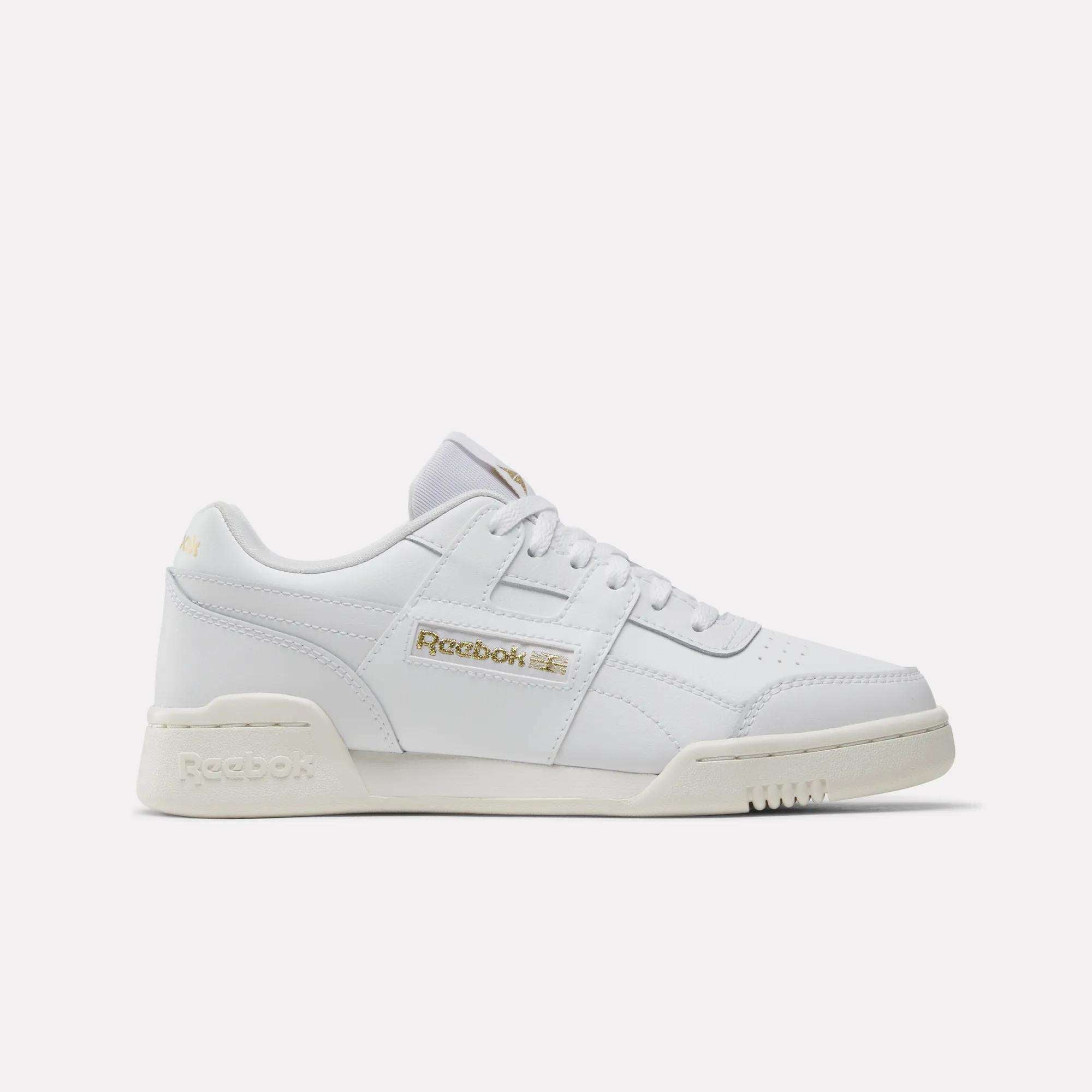 The Reebok Workout Plus Shoes - Big Kids are white low-top sneakers with gold branding on the side and heel, white laces, and an off-white sole—ideal sporty footwear for juniors, shown on a plain white background.