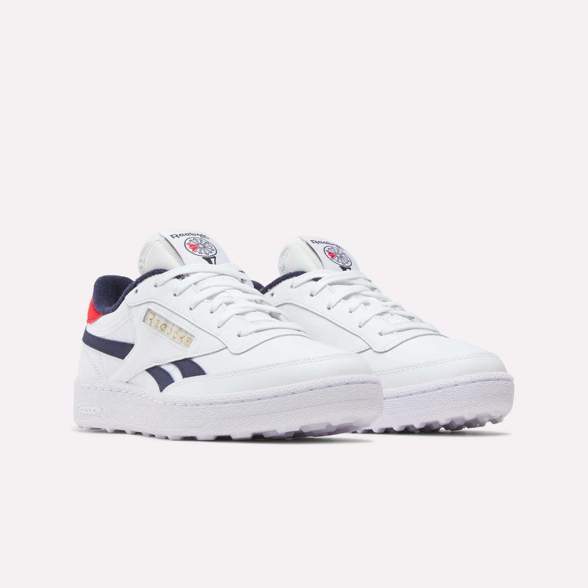 The Men's Club C Revenge Golf Shoes by Reebok are white with navy blue and red accents, feature the Reebok logo on the tongue and side, and have a spikeless outsole for traction, shown against a plain light background.