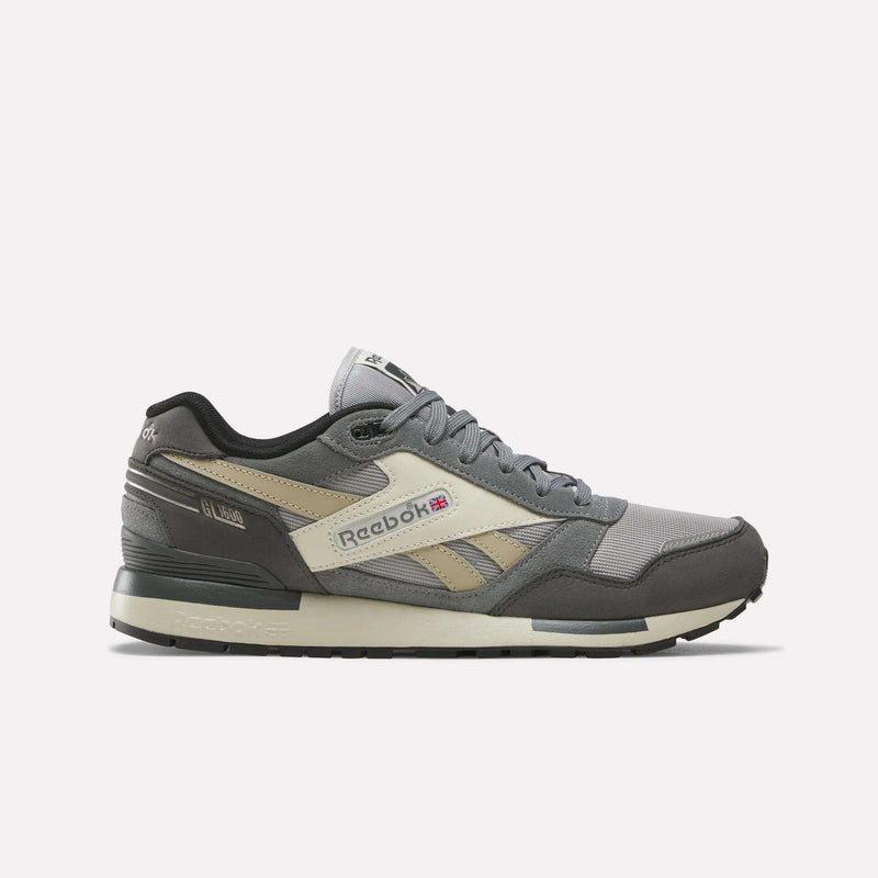 A Reebok Men's GL1600 Shoe in Almostblack/Grey4/Linen, featuring black accents, a white midsole, and a gray outsole is displayed in side profile against a plain white background. This stylish sneaker boasts premium suede construction with subtle reflective details for enhanced visibility.