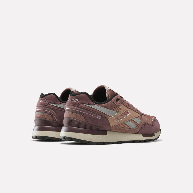 A pair of Reebok Men’s GL1600 Shoes in mauve and beige suede are displayed angled from the back and side on a plain white background, showcasing the Reebok logo and retro-inspired design. Subtle reflective elements bring a modern update to these classic sneakers.