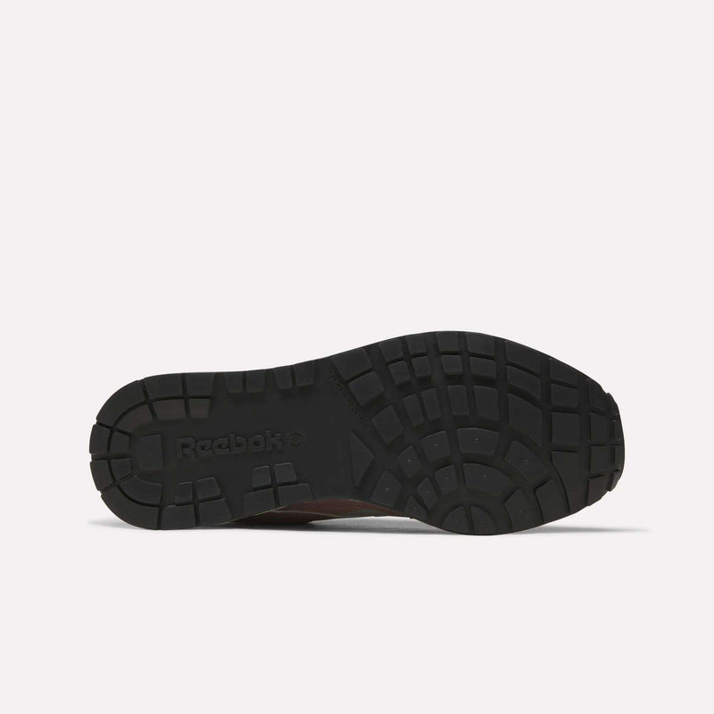 The image displays the bottom view of a Reebok Men’s GL1600 Shoe sole in black, featuring a textured tread pattern distinctive to this series. The sole showcases the Reebok logo on the left side, underscoring the meticulous design typical of these classic sneakers.