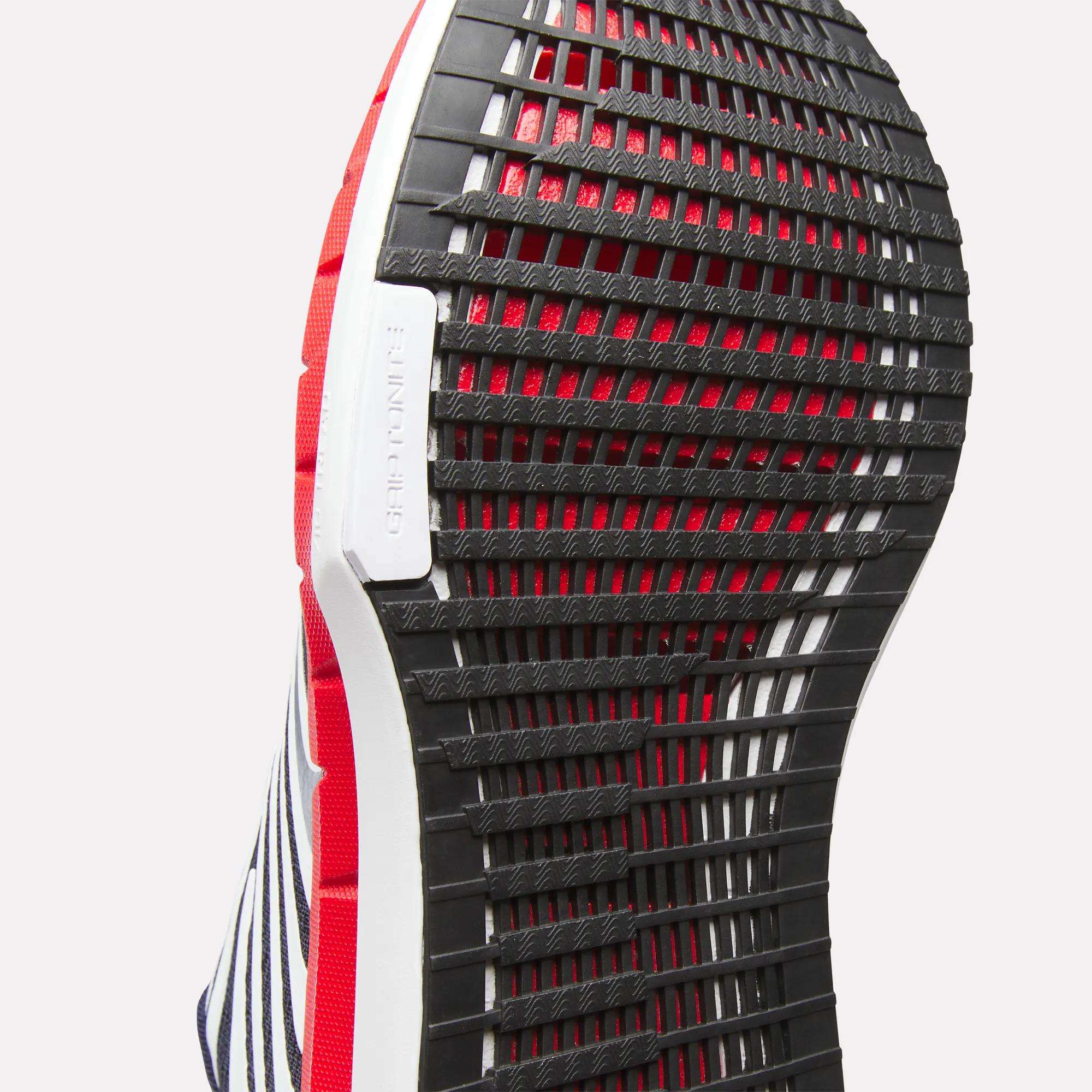 Close-up of the black, grid-patterned rubber outsole on Reebok Men's FloatZig Tread Running Shoes, accented in red, white, and navy blue, with a visible GRIPTONNER label—designed for optimal treadmill workouts.