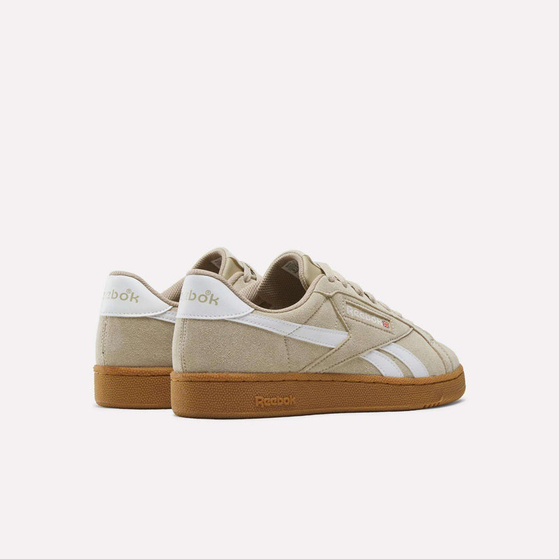 A pair of Reebok Men’s Club C Grounds UK 1.0 shoes in beige suede, featuring white stripes and tan rubber soles, is displayed from a rear side angle on a plain light background, showcasing classic Reebok heritage style.