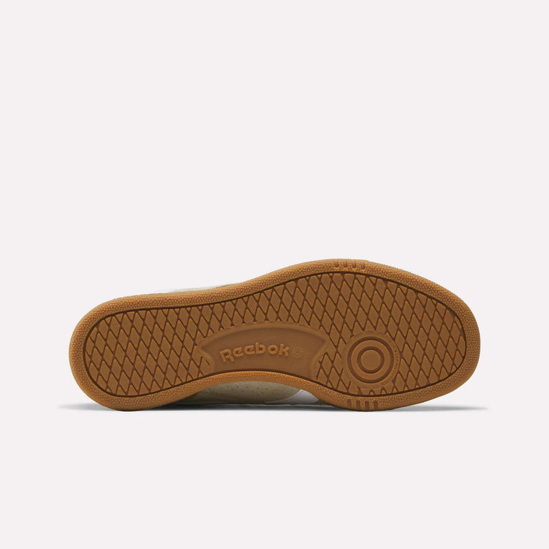 The image shows the brown rubber sole of a Reebok Men’s Club C Grounds UK 1.0 shoe, featuring a geometric tread pattern with the Reebok logo embossed in the center, embodying classic Reebok heritage.