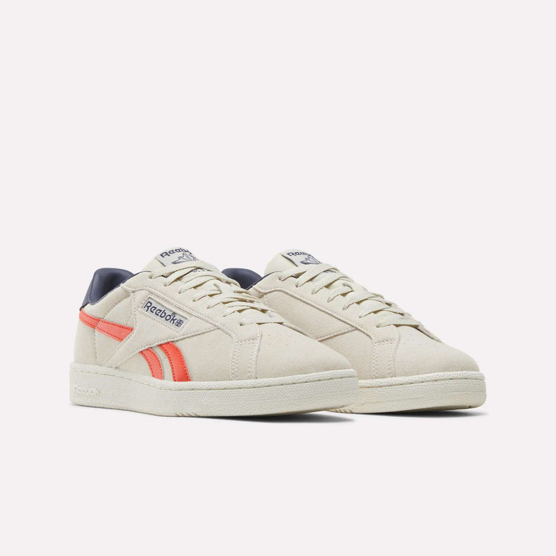 A pair of Men’s Club C Grounds UK 1.0 Shoes by Reebok in beige, featuring coral and navy accents, displayed on a white background. These men’s sneakers showcase lace-up fronts, classic retro styling, and premium suede accents that reflect their authentic Reebok heritage.