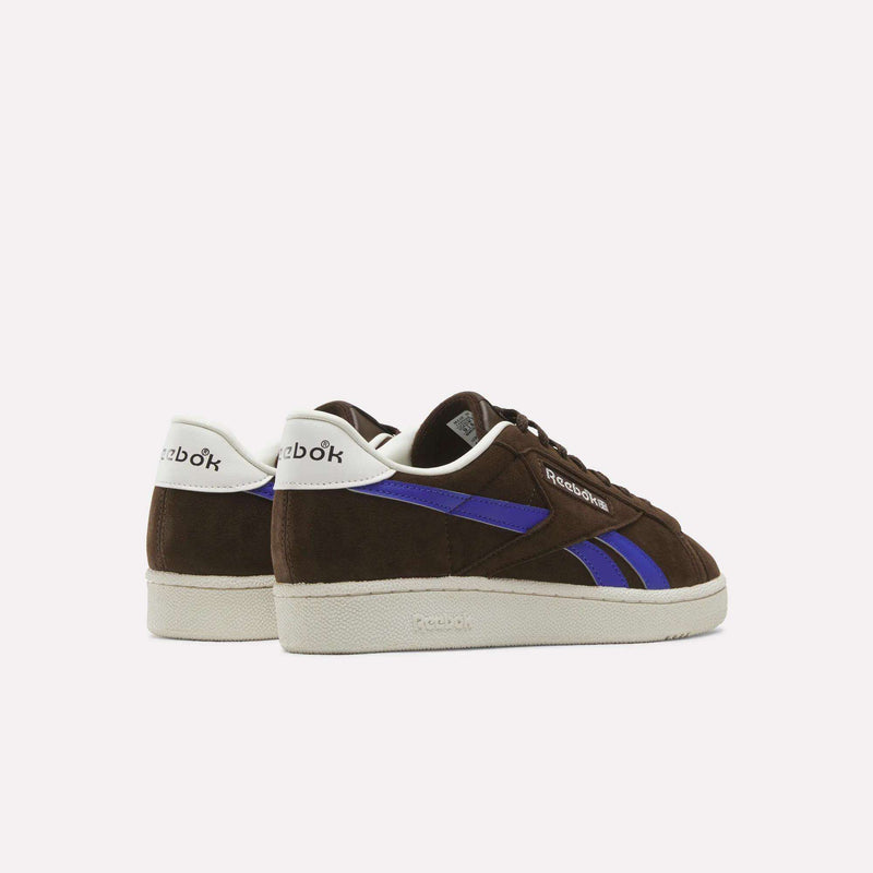 A pair of Reebok Men’s Club C Grounds UK 1.0 Shoes in brown suede with blue stripes and white soles, displayed from the back and side on a plain background—these sneakers capture a timeless, vintage-inspired style.