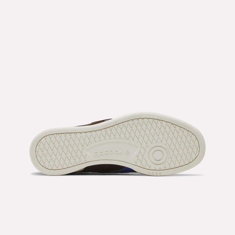 The image displays the white rubber sole of the Reebok Men’s Club C Grounds UK 1.0 Shoes, viewed from below. It features a herringbone tread pattern and the Reebok logo at the center. This classic design is characteristic of both vintage and suede sneakers, emphasizing Reebok’s enduring style.