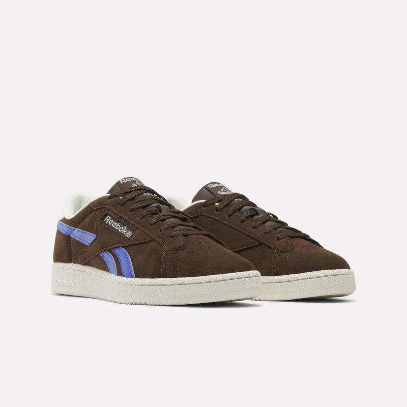 A pair of Reebok Men’s Club C Grounds UK 1.0 Shoes made of brown suede, featuring purple side stripes and white soles, are placed side by side on a plain white background.