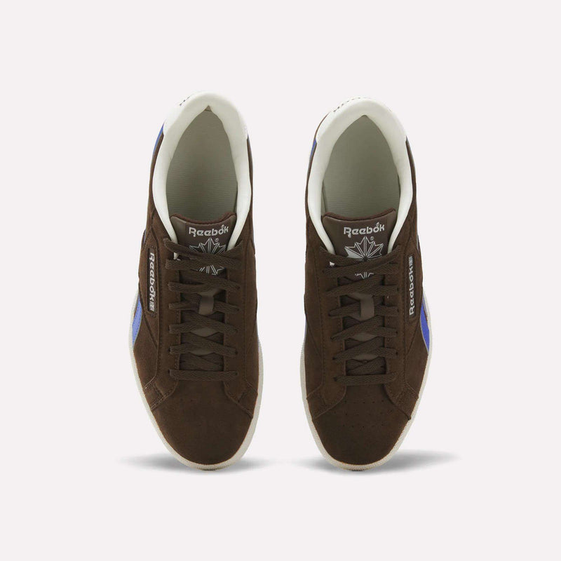 A pair of Men’s Club C Grounds UK 1.0 Shoes by Reebok in brown suede, featuring white soles and blue stripes, are seen from above against a plain white background, highlighting their classic vintage sneaker style.