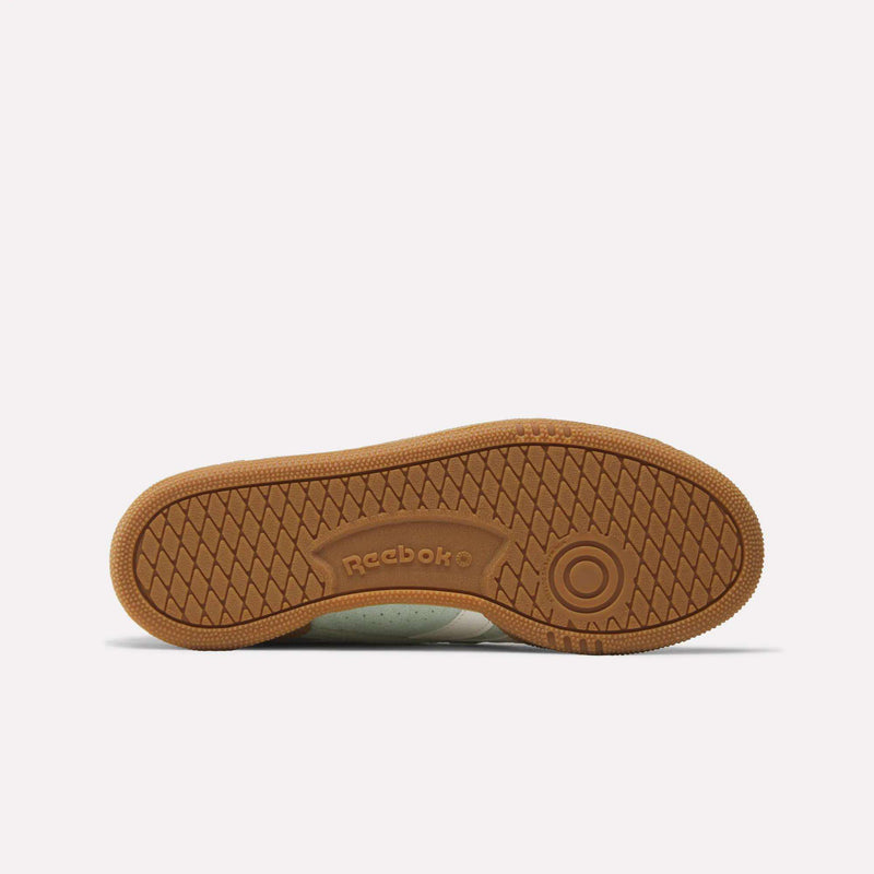 Bottom view of the Reebok Women’s Club C Grounds UK 1.0 Shoes featuring a brown rubber sole with a circular pattern near the heel, herringbone tread, and the Reebok logo embossed at the center. The plain white background highlights the classic appeal of these vintage-inspired sneakers.