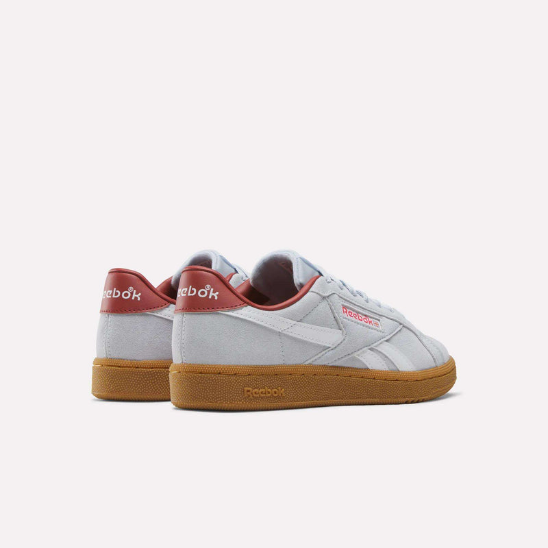 A pair of Reebok Women’s Club C Grounds UK 1.0 Shoes featuring white uppers, gum soles, and red accents on the heel and logo are displayed on a plain white background. These classic sneakers combine timeless style with everyday comfort for women.