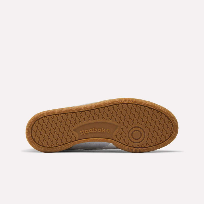 The image shows the gum-colored sole of the Reebok Women’s Club C Grounds UK 1.0 Shoes, featuring a herringbone tread pattern and the Reebok logo in the center—a detail that reflects classic heritage often seen in their suede sneaker designs.