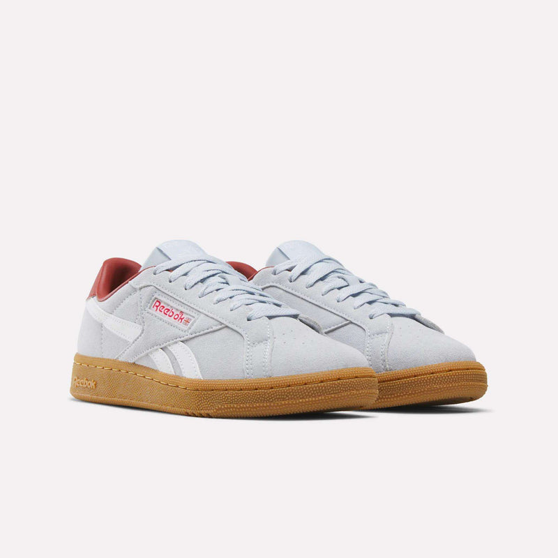 A pair of Women’s Club C Grounds UK 1.0 Shoes by Reebok in light gray suede, featuring gum-colored soles, white side stripes, red Reebok branding, and red interior lining, displayed on a plain white background.