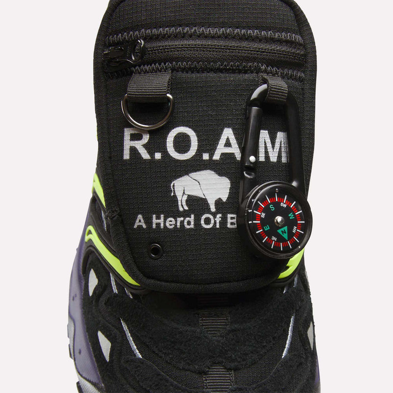 Close-up of a Reebok x A Herd of Bison Men’s Trailzillo DMX shoe tongue with a zippered pocket featuring the text R.O.A.M and a bison icon, with a small compass attached by a carabiner.