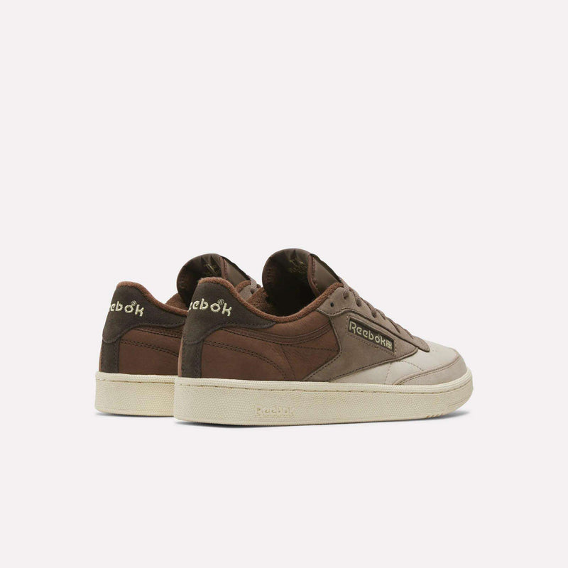 A pair of Reebok Men’s Club C 85 Shoes in brown and beige are displayed on a plain white background, angled to show the side and back, with the Reebok logo visible on both the heel and side.