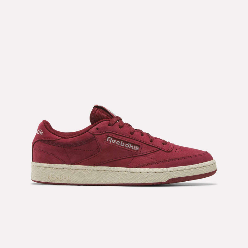 A Reebok Men’s Club C 85 Shoe in Classicburgundy with a linen-hued sole is displayed on a plain white background. This stylish tennis sneaker features the Reebok logo and text in English and Asian characters on the side, crafted from premium nubuck leather for a classic look and feel.