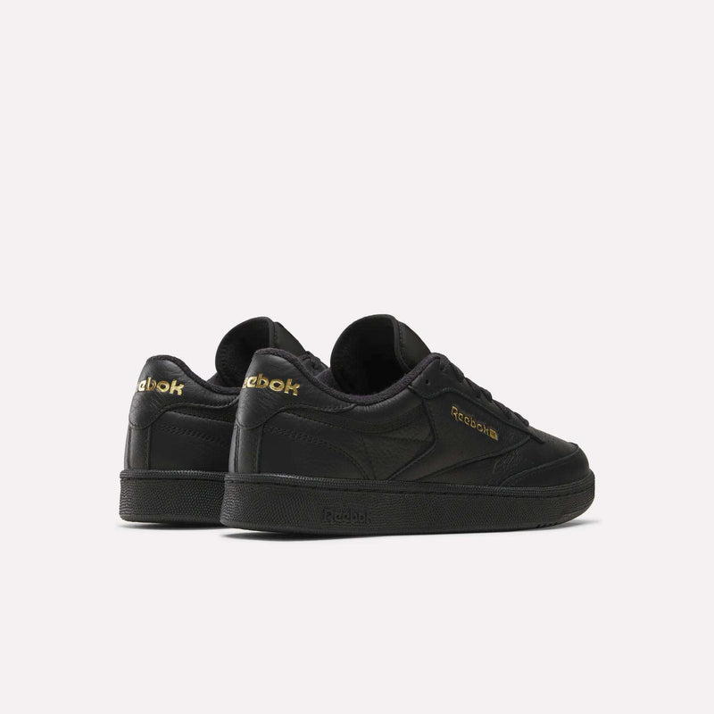 A pair of Men’s Club C 85 Shoes by Reebok in black, crafted from premium garment leather with gold Reebok branding, is displayed on a plain white background. The retro sneakers are angled to showcase both the side and back views.