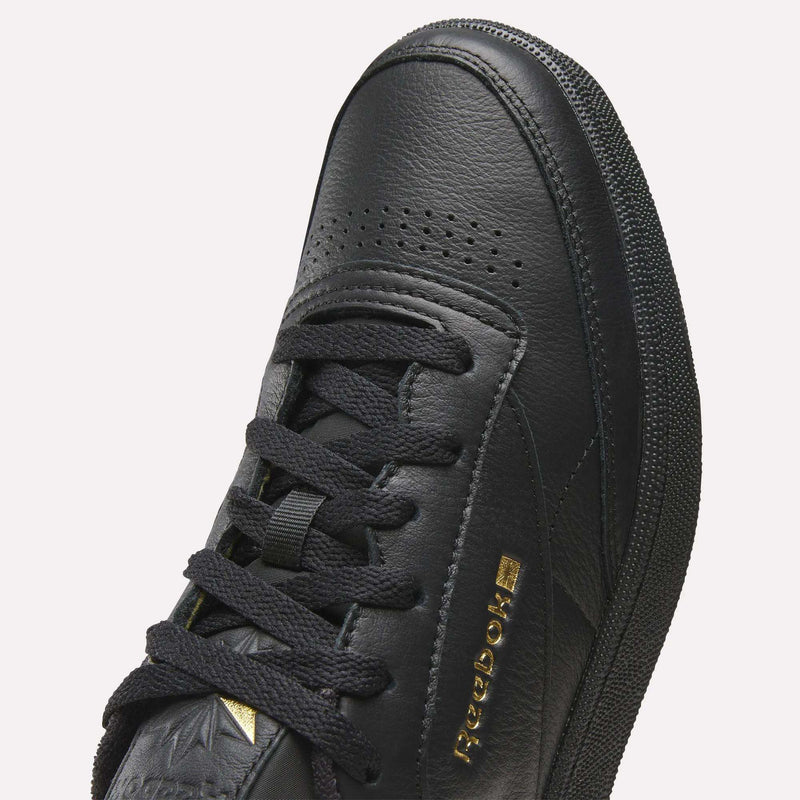 Close-up of the Reebok Men’s Club C 85 Shoes in black, showcasing premium garment leather, black laces, perforated toe, textured side panels, and gold Reebok branding on the side.
