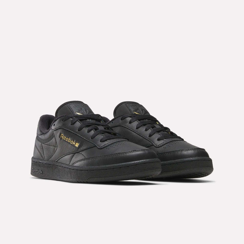 A pair of Reebok Men’s Club C 85 shoes in black, showcasing gold branding, crafted from premium garment leather. These low-top sneakers feature matching black soles and laces, and are presented at a slight angle to display both the outer side and front—a modern take on the iconic Reebok classic.