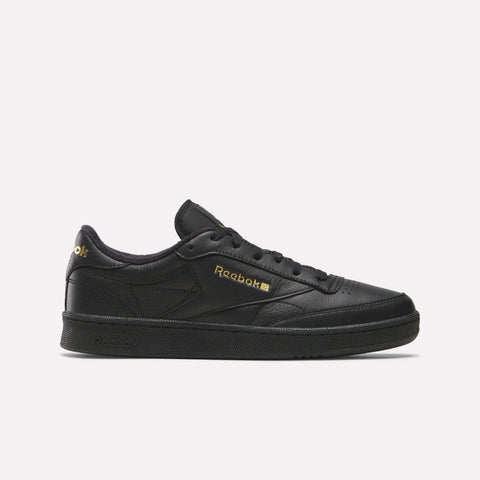 A side profile of the Men’s Club C 85 Shoes by Reebok is shown against a plain white background. This sneaker, in a Black/Almostblack/Gold colorway, is crafted from premium garment leather and features gold Reebok branding. The iconic design and elevated materials capture the timeless appeal of retro shoes.