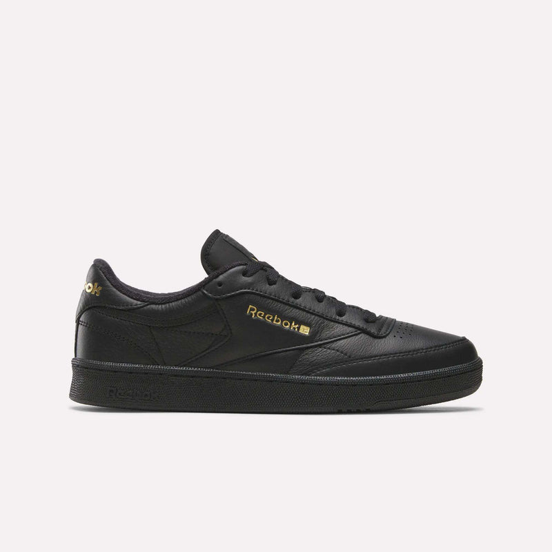 A side profile of the Men’s Club C 85 Shoes by Reebok is shown against a plain white background. This sneaker, in a Black/Almostblack/Gold colorway, is crafted from premium garment leather and features gold Reebok branding. The iconic design and elevated materials capture the timeless appeal of retro shoes.