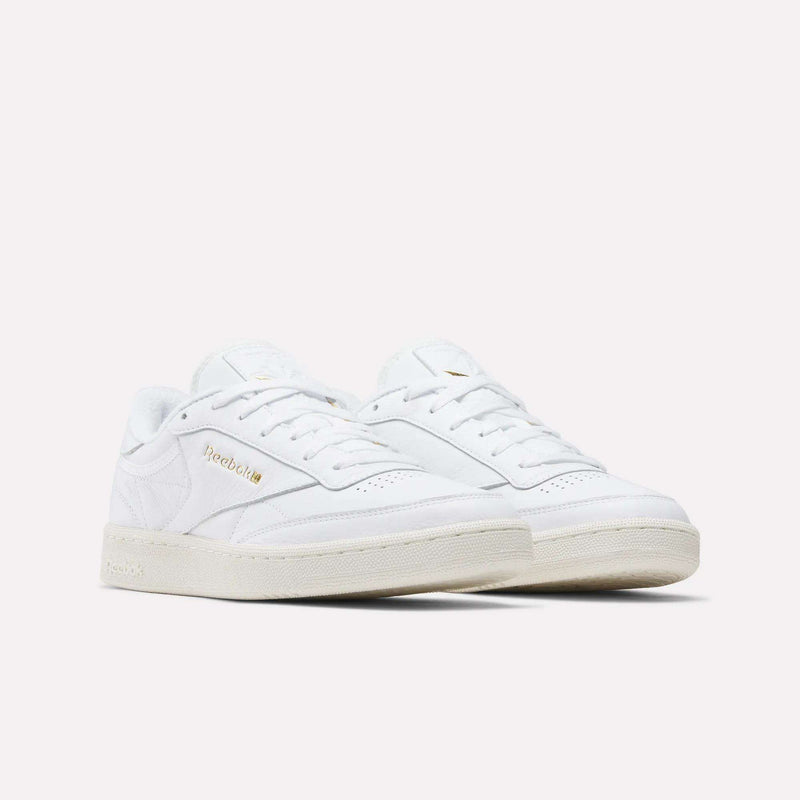 The Reebok Men's Club C 85 Shoes feature white premium garment leather, off-white soles, gold Reebok branding on the side, and a classic low-top silhouette. Displayed against a plain white background.