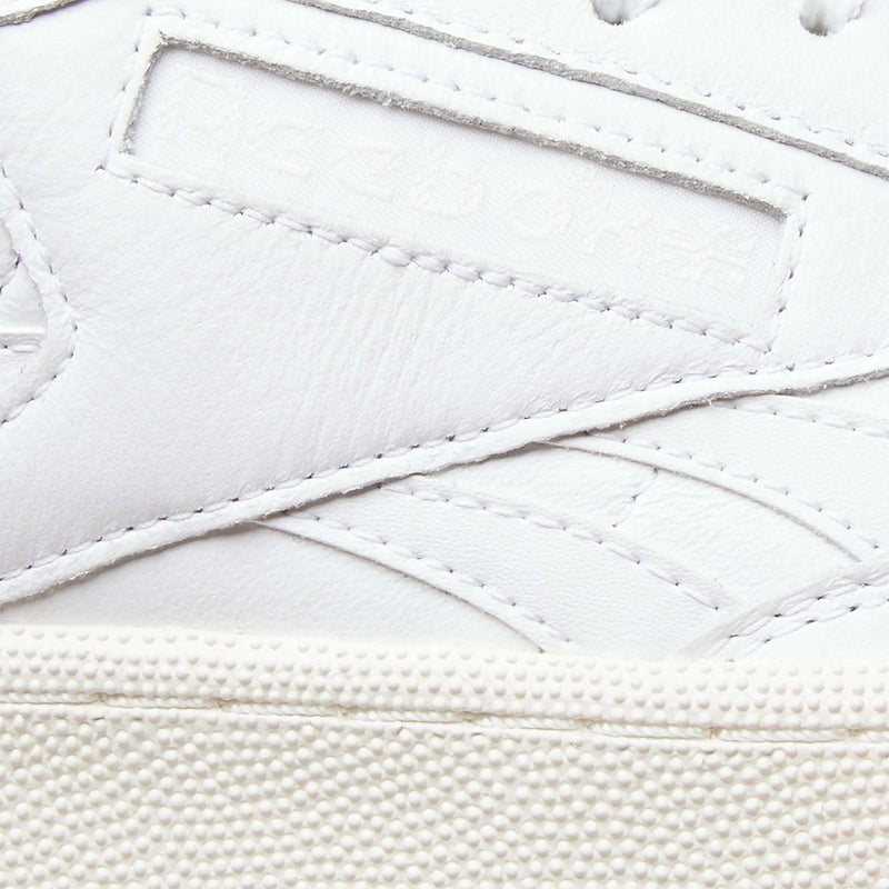 Close-up of the Reebok Men’s Club C Revenge Shoes in white leather, featuring visible stitching and embossed Reebok branding on the side. The textured rubber sole is shown at the bottom of the image, emphasizing its tennis-inspired sneaker design.