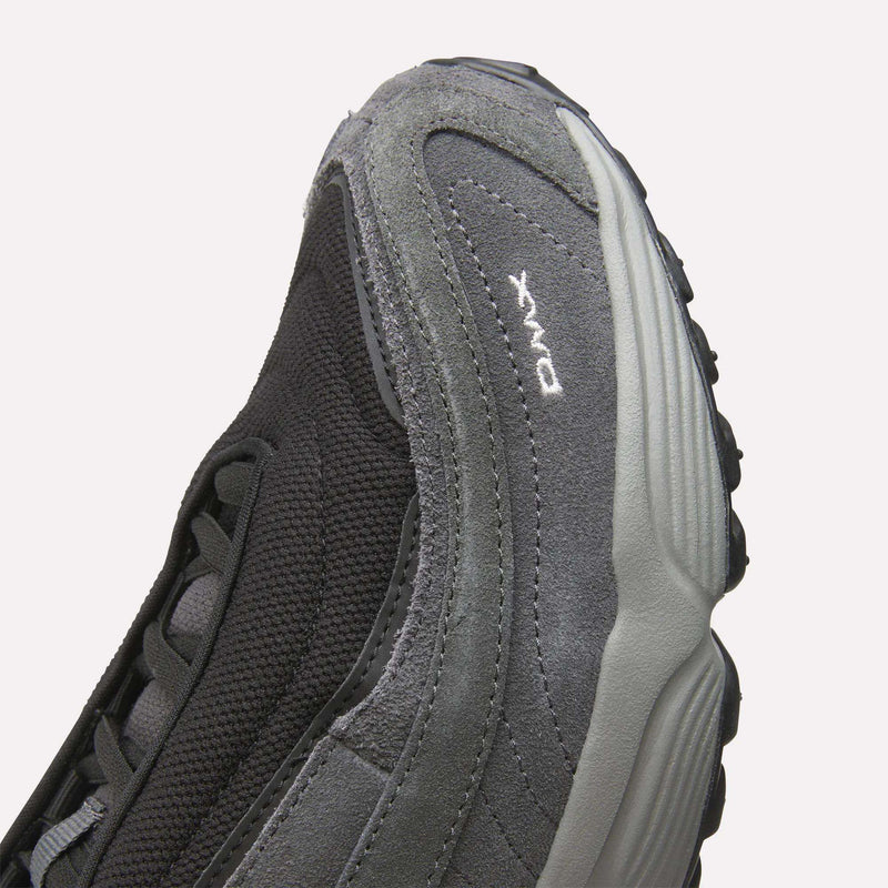 Close-up of a gray and black Men’s Daytona DMX Shoe by Reebok, highlighting its textured suede and mesh upper, intricate stitching details, and “DMX” embroidered in white near the sole. This Motorsport-inspired sneaker offers air cushioning for superior comfort and support.