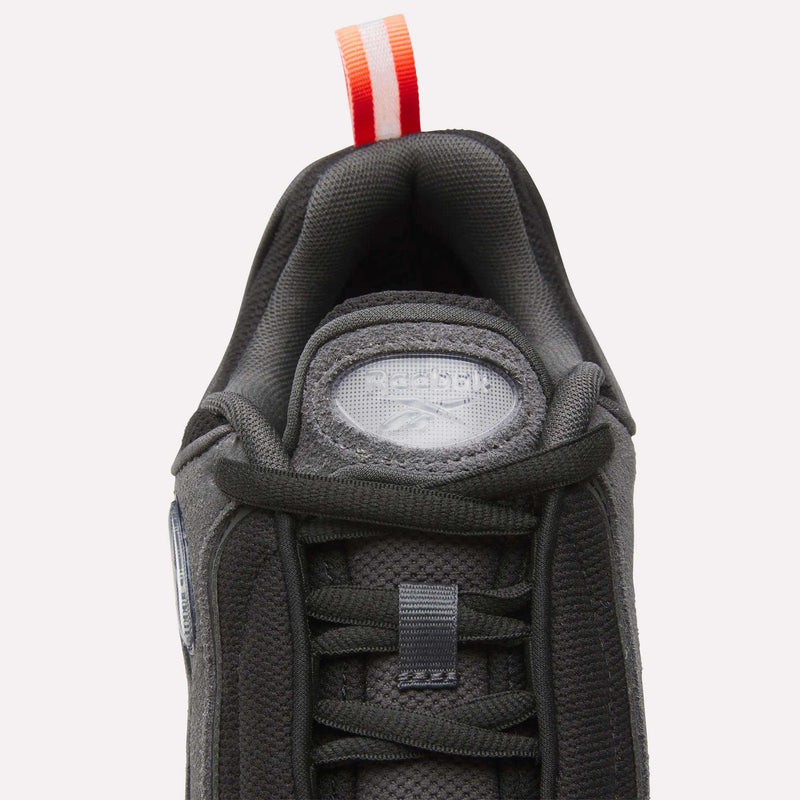 Top view of a dark gray Reebok Men’s Daytona DMX Shoe, featuring a textured fabric upper, thick padded collar, and a red-and-white striped pull tab on the heel. This Motorsport-inspired sneaker offers air cushioning for comfort and showcases a tongue with a circular label accented by a small silver emblem and text.