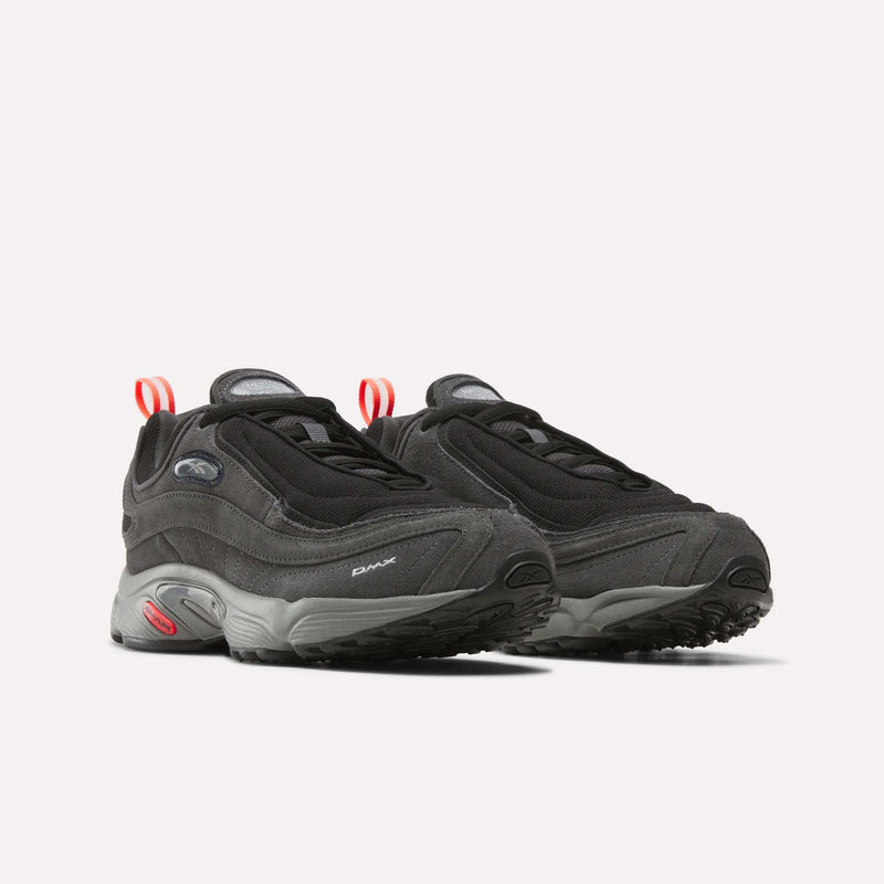 A pair of Reebok Men’s Daytona DMX Shoes in black and gray, featuring chunky soles, red heel pulls, air cushioning, and mesh detailing, displayed on a plain white background.