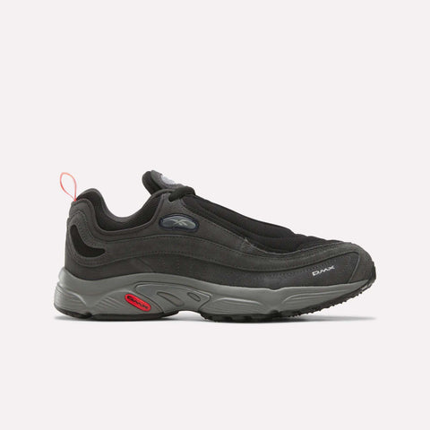A side profile view of the Reebok Men’s Daytona DMX Shoes in Black/Almostblack/Neoncherry displays a black athletic sneaker with a thick gray air-cushioned sole, mesh upper, and a red (neon cherry) pull tab at the heel, set against a plain white background—these motorsport-inspired sneakers combine distinctive style and high performance.