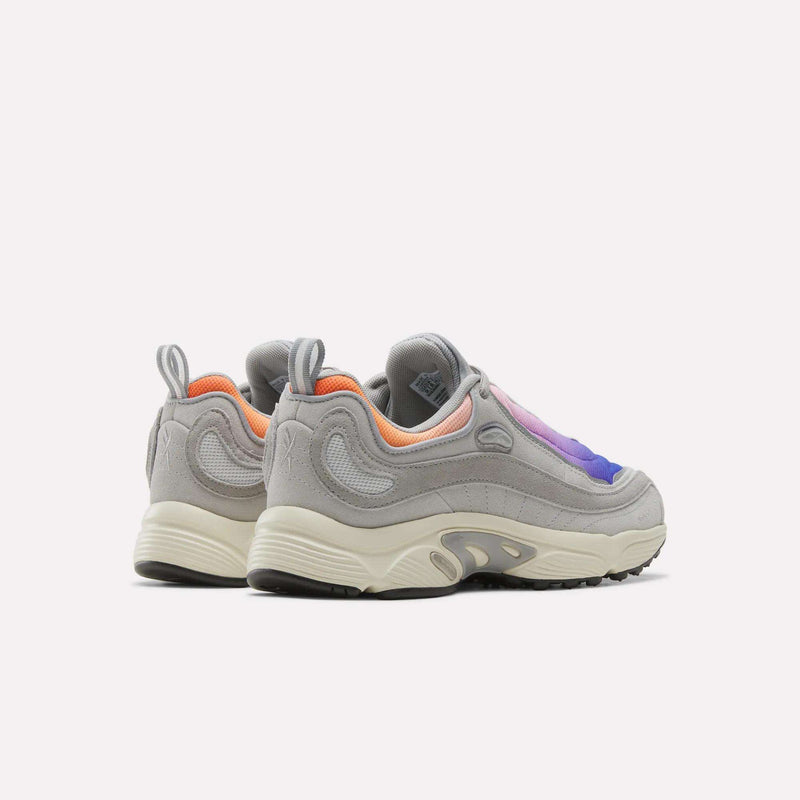 A pair of Men’s Daytona DMX Shoes by Reebok in gray, featuring white soles, orange inner lining, and gradient purple and blue accents with motorsport-inspired design details and advanced air cushioning, displayed at an angle on a plain white background.