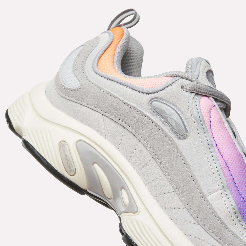 A close-up of the Reebok Men’s Daytona DMX Shoes featuring light gray suede, pastel orange and purple accents, a white midsole, and black outsole against a plain background—these sneakers showcase Motorsport-inspired design with advanced DMX air cushioning from Reebok for superior comfort.