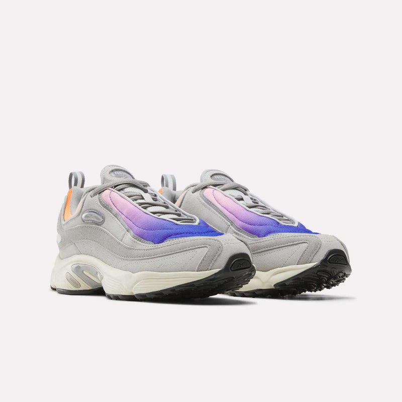 A pair of Men’s Daytona DMX Shoes by Reebok, designed in gray with white soles, blue and purple gradient panels, black outsoles, and subtle orange accents on the heels—these Motorsport-inspired sneakers also feature air cushioning for enhanced comfort—all showcased against a plain white background.