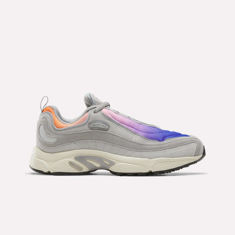 A men’s sneaker from Reebok, the Daytona DMX Shoes, in New Grey 1 with Breakout Blue and Expressive Pink accents, features mesh and suede panels, a thick off-white sole with air cushioning, and black tread. Displayed against a plain white background, this design pays homage to the Daytona DMX’s motorsport-inspired heritage.