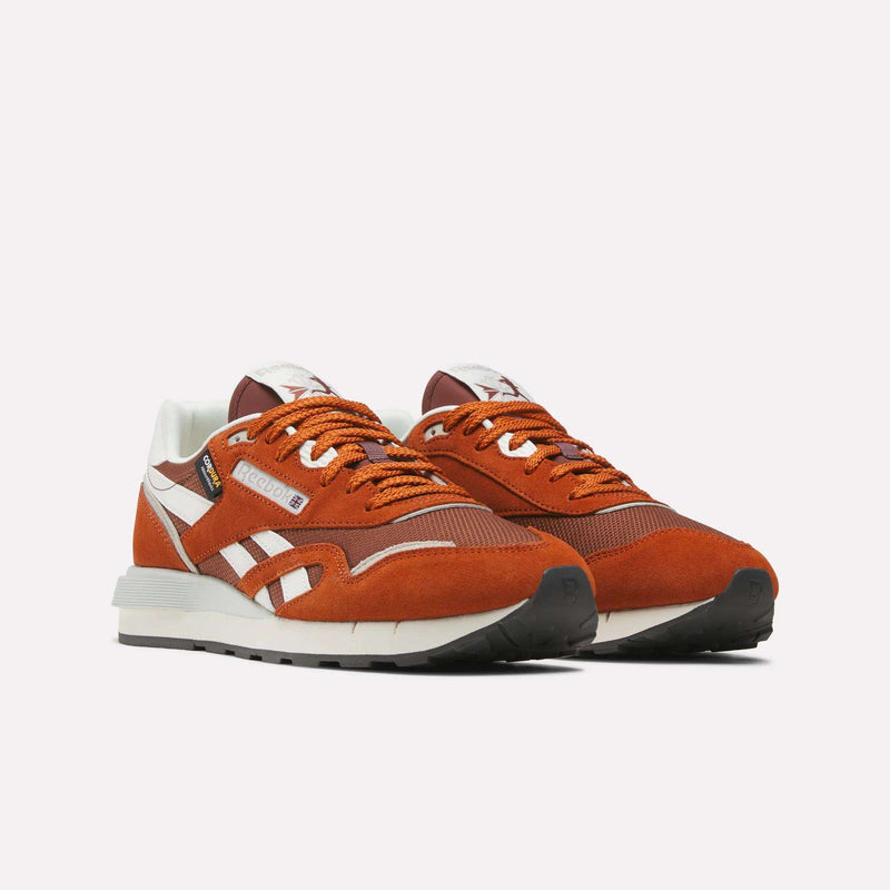 A pair of Reebok Men’s Classic Nylon 89 Shoes in orange and white, crafted from mixed materials with white soles, orange laces, black accents, and a comfortable EVA midsole, are positioned side by side on a plain white background.