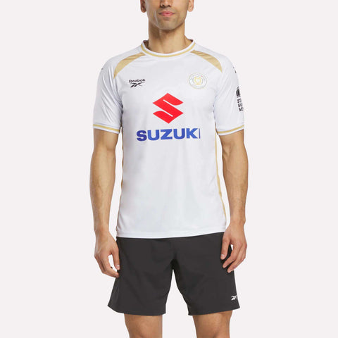 A man wears the Reebok Men's MK Dons Home Jersey—white with gold accents and the “SUZUKI” logo—paired with black athletic shorts, standing against a plain background. This football jersey is inspired by Milton Keynes’ pride.