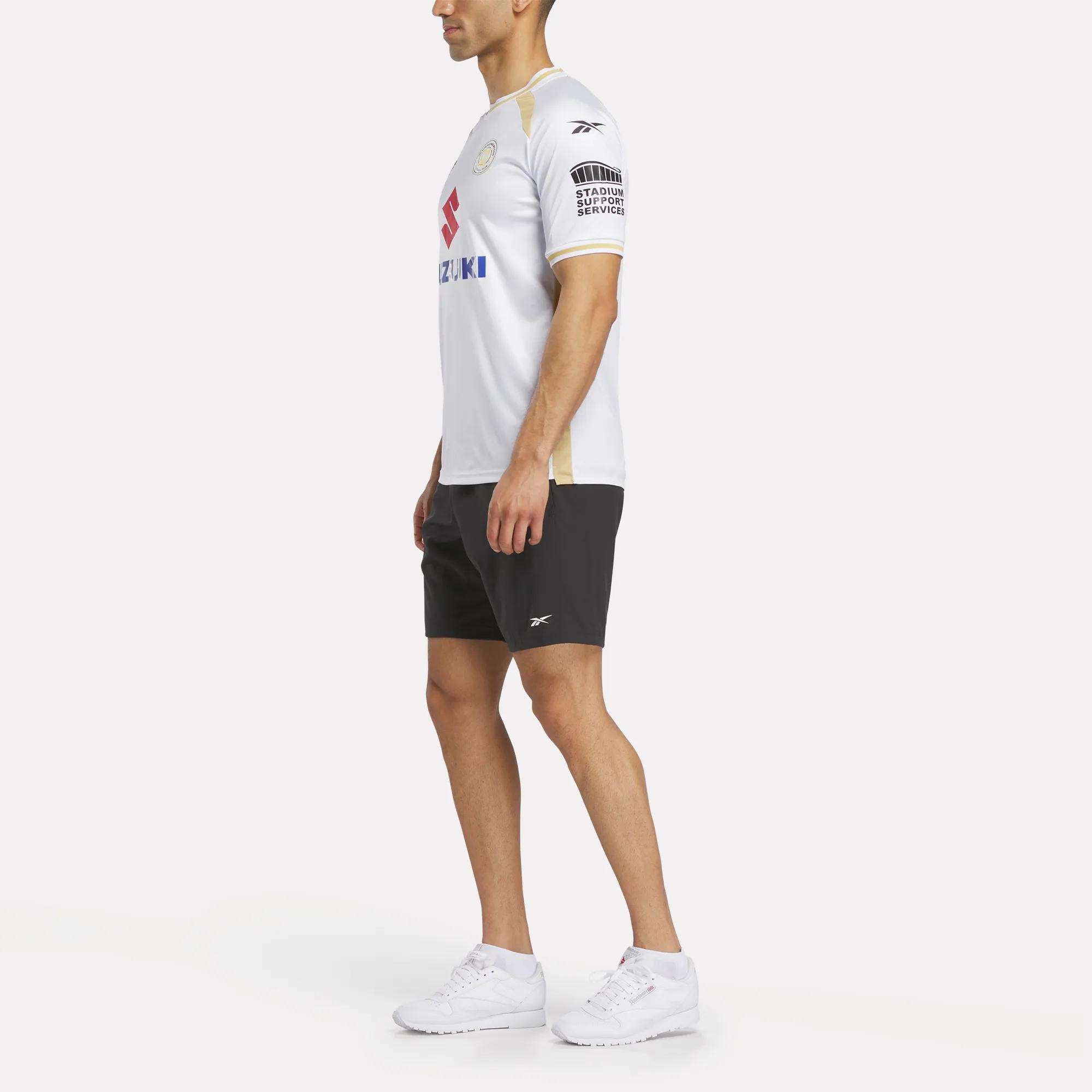 A man stands sideways against a white background wearing the Reebok Men's MK Dons Home Jersey, paired with black shorts and white sneakers.