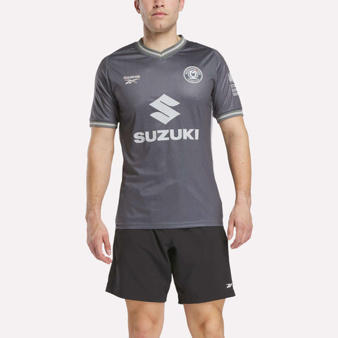A man wears a Reebok Men's MK Dons Away Jersey featuring SUZUKI and logos, paired with black athletic shorts, standing against a plain light background.