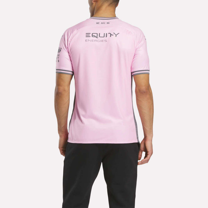 A person in Milton Keynes faces away, wearing a Reebok Men's MK Dons Third Jersey in light pink with gray trim and "EQUITY ENERGIES" on the back, paired with black pants.