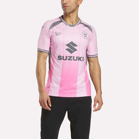 A man wears the Men's MK Dons Third Jersey by Reebok, showing pink and light gray stripes with Suzuki and Reebok logos, paired with black athletic pants, against a plain white background.