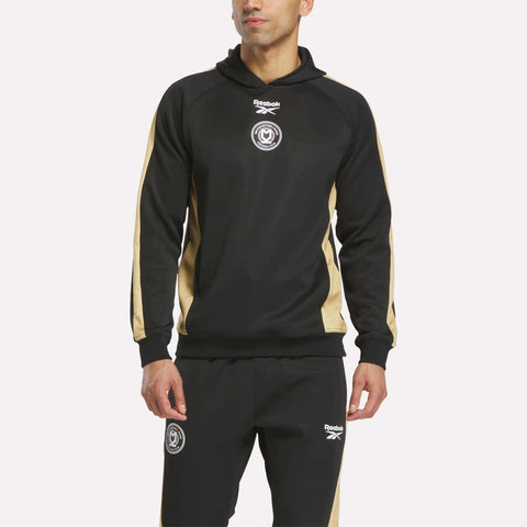 A man in a black Reebok tracksuit with beige stripes and logos on the chest and thigh stands against a white background, ideal for pairing with the Reebok Men's MK Dons Travel Hoodie.