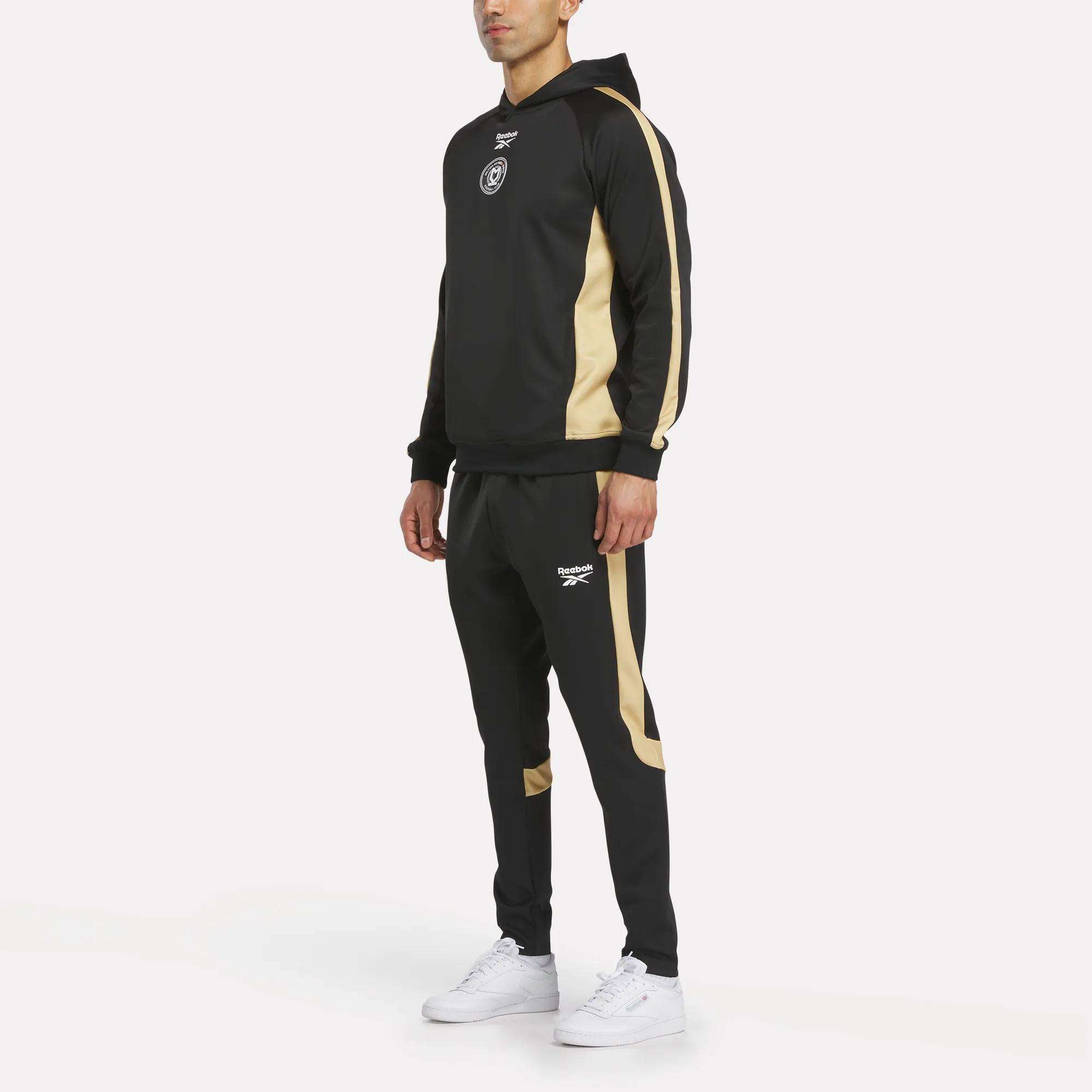 A man wears the Reebok Men's MK Dons Travel Hoodie tracksuit in black with gold stripes on the sleeves and legs, a hood, and a white Reebok logo, paired with white sneakers. The background is plain white.