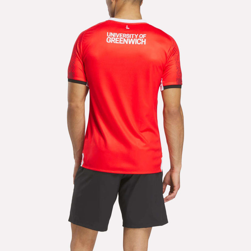 A person faces away, wearing the Reebok Men's Charlton Home Jersey in bright red with club badge details and black athletic shorts, set against a plain white background.