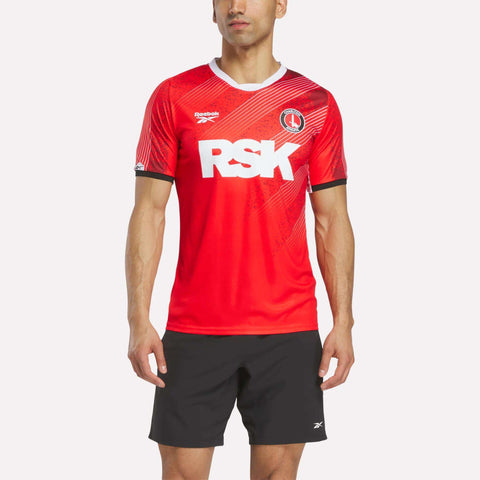 A person wearing a red Reebok Men's Charlton Home Jersey with RSK in white letters and black shorts stands against a plain white background. The jersey features a round collar, club badge design, and club crest on the left chest.