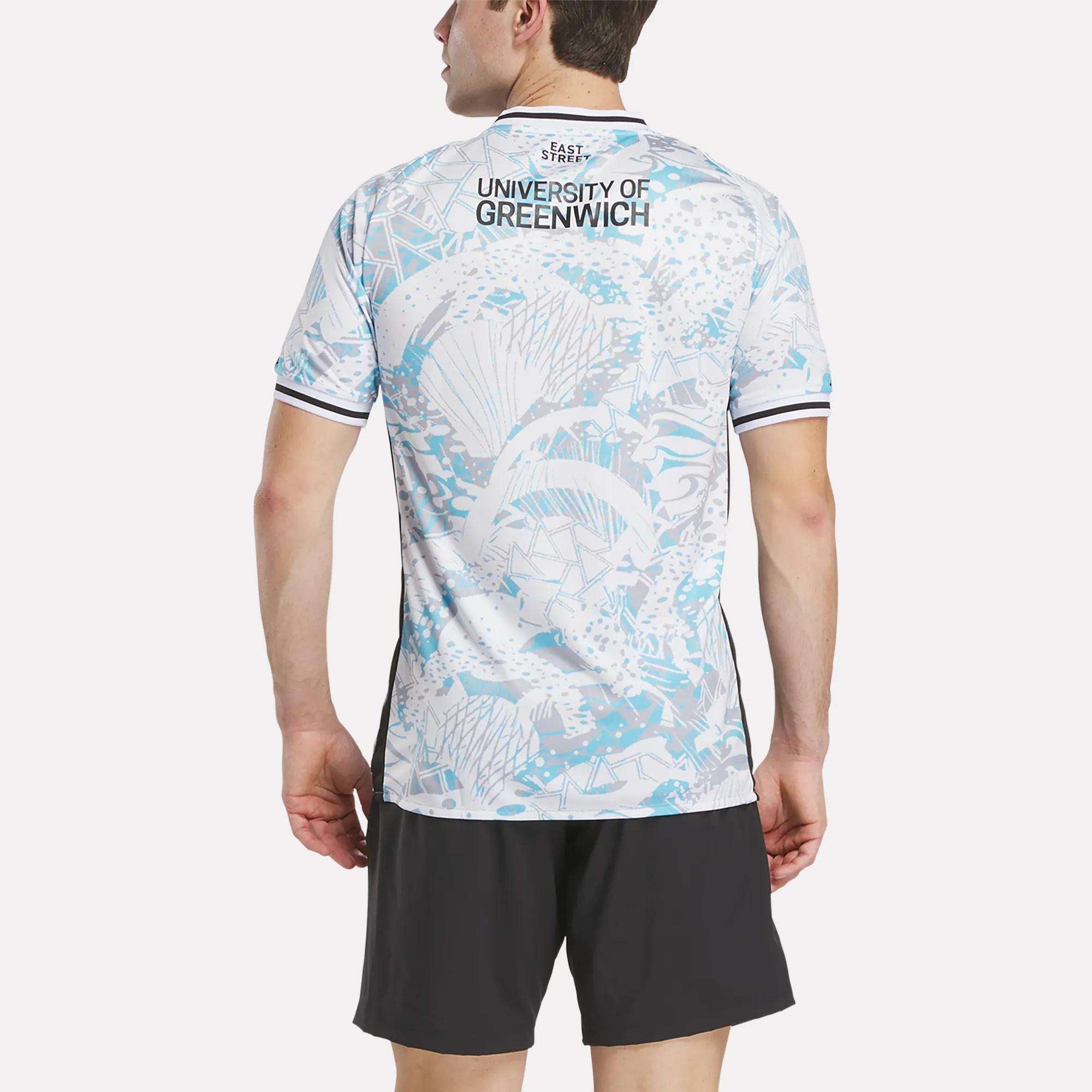 A man faces away wearing the Reebok Men's Charlton Away Jersey, featuring an aquatic blue and white heritage-inspired design with "University of Greenwich" on the back, paired with black athletic shorts.